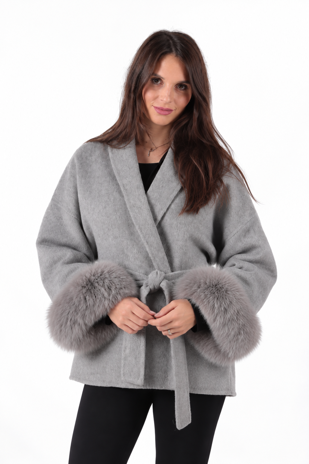 A woman wearing a light gray wrap-style coat with plush gray fox-fur trimmed cuffs and a matching tie belt, paired with black pants, against a plain white background.