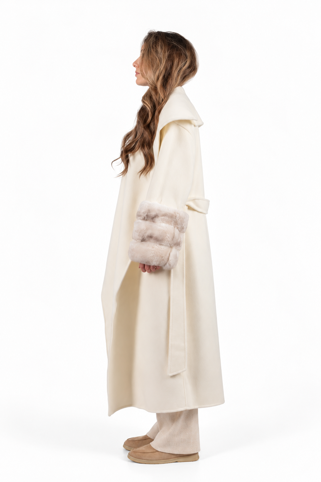 A woman shown in profile wearing a long cream-colored coat with a soft, fuzzy light beige trim at the cuffs, paired with matching wide-leg trousers and beige slippers, against a plain white studio background.