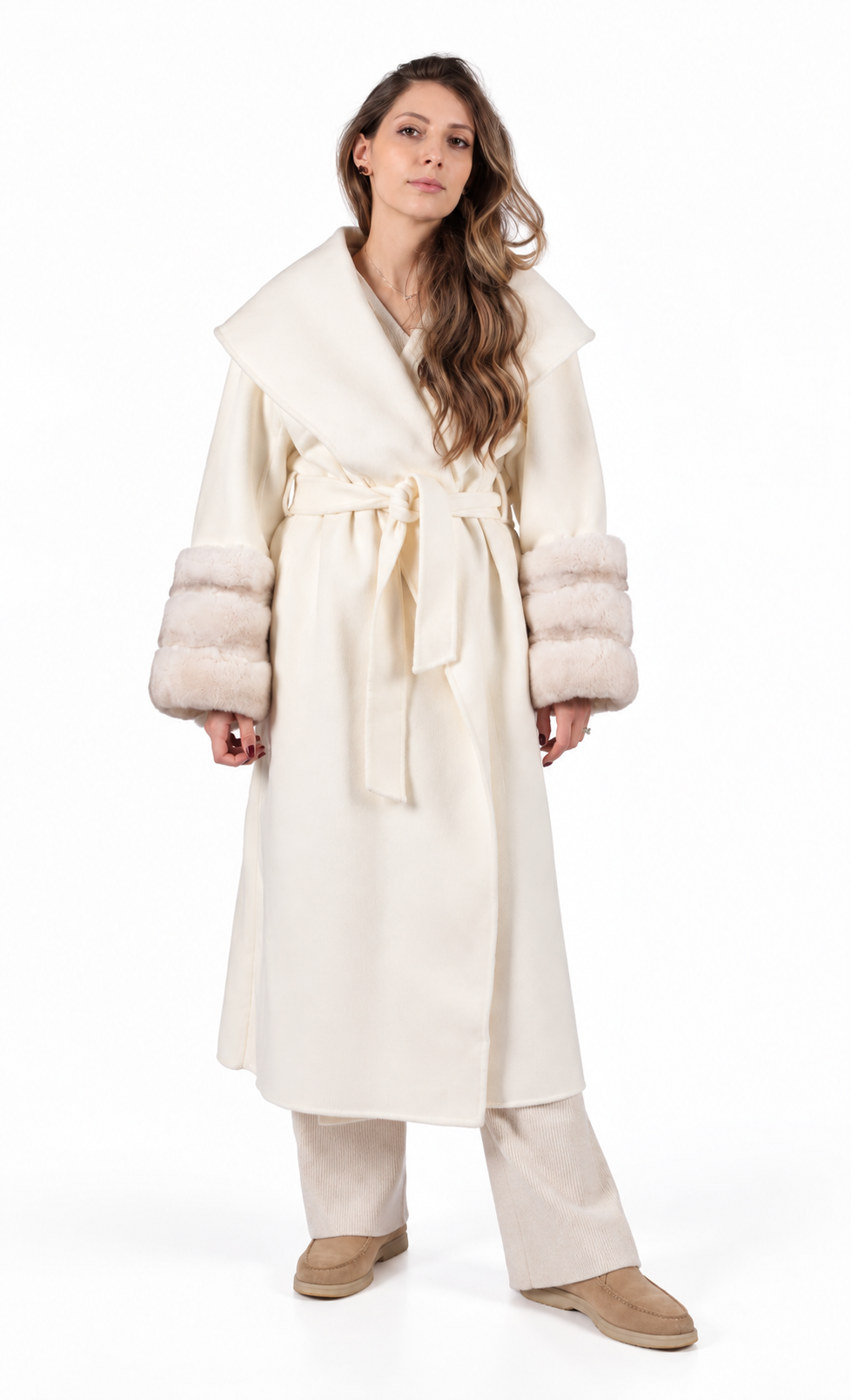 A woman modeling a long cream-colored coat with a wide collar, matching waist tie, and fluffy rabbit-fur trimmed cuffs, paired with beige pants and tan shoes against a plain white background.