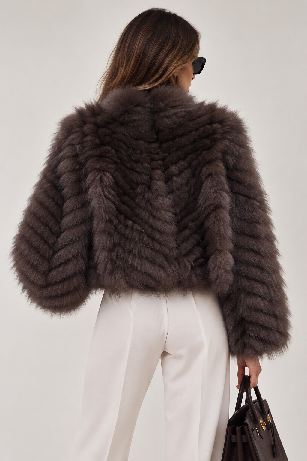 A woman shown from the back wearing a cropped, dark brown shaggy fox fur jacket, white trousers, and holding a black handbag against a plain light gray background.