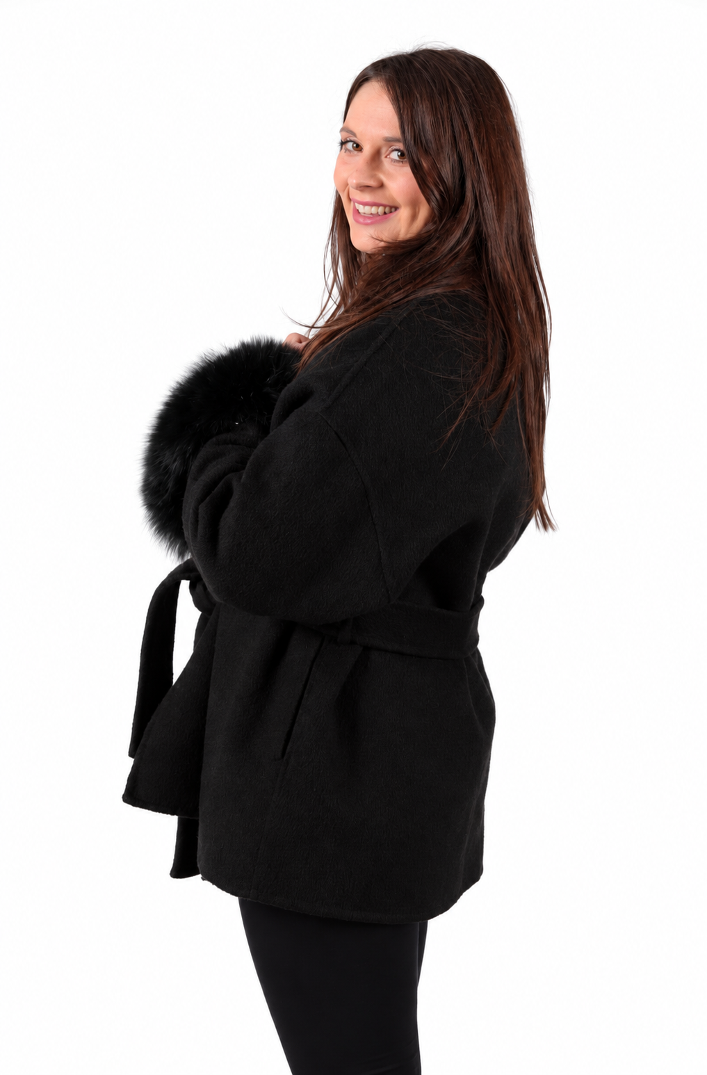 A woman with long brown hair is shown from the side, wearing a black belted coat with a black fur detachable cuffs photographed against a solid white background.